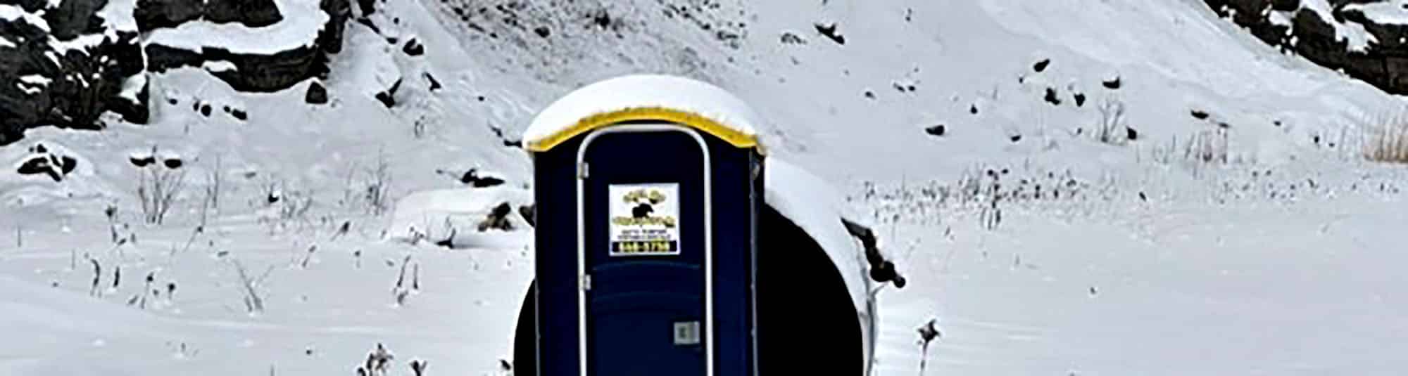 Portable toilet covered in snow on a snowy landscape with rocky background