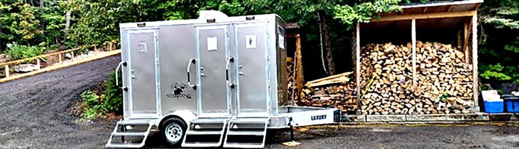 Portable washroom trailer near stacked firewood in a forest setting